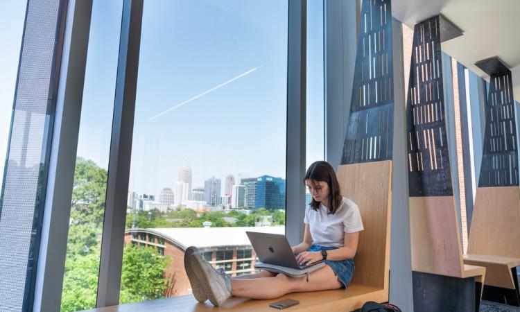 A woman sits with a laptop next to a large window