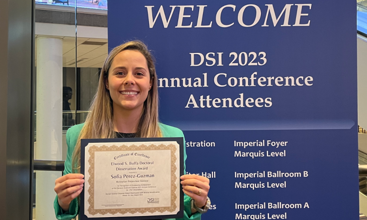 A woman holds a certificate in front a conference welcome sign 