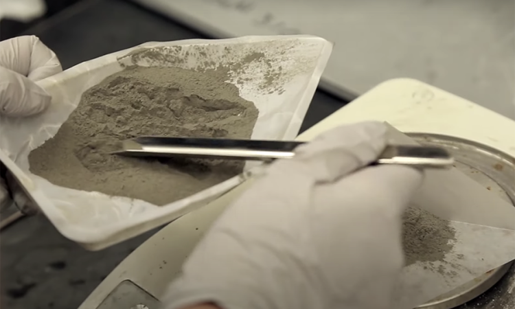 A close up of coal ash and a silver instrument in a dish held by hands in white latex gloves 