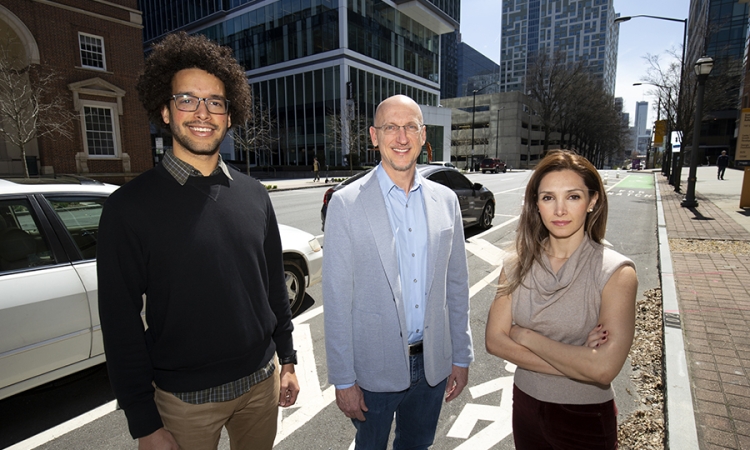 Two men and one woman stand together on the street in front of cars and buildings