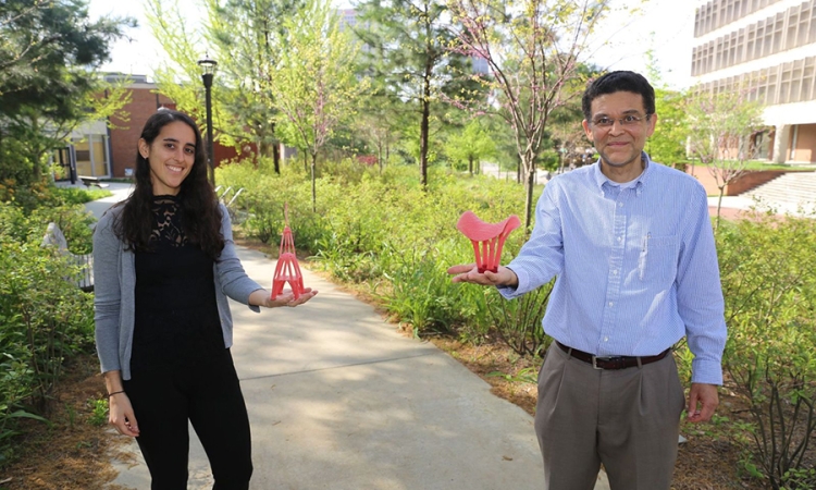 PhD Student Emily Sanders, left, and Professor Glaucio Paulino, right, hold red 3-D printed models as they stand on a campus sidewalk surrounded by greenery. 