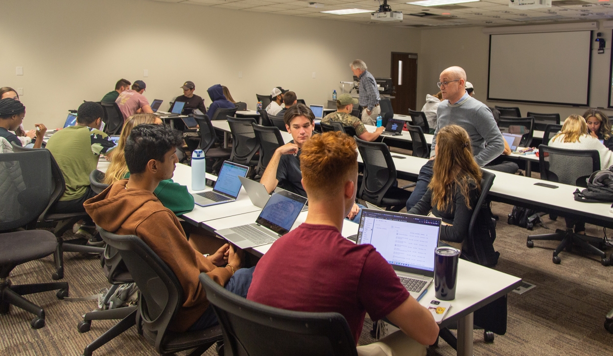 Two instructors walk around a classroom talking to seated groups of students with laptops.