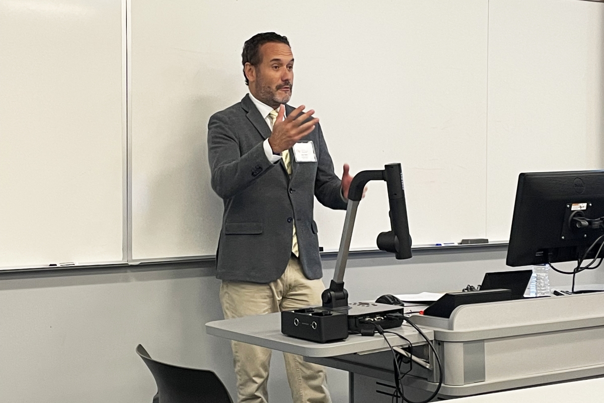 A man in a blazer stands in front of a white board 