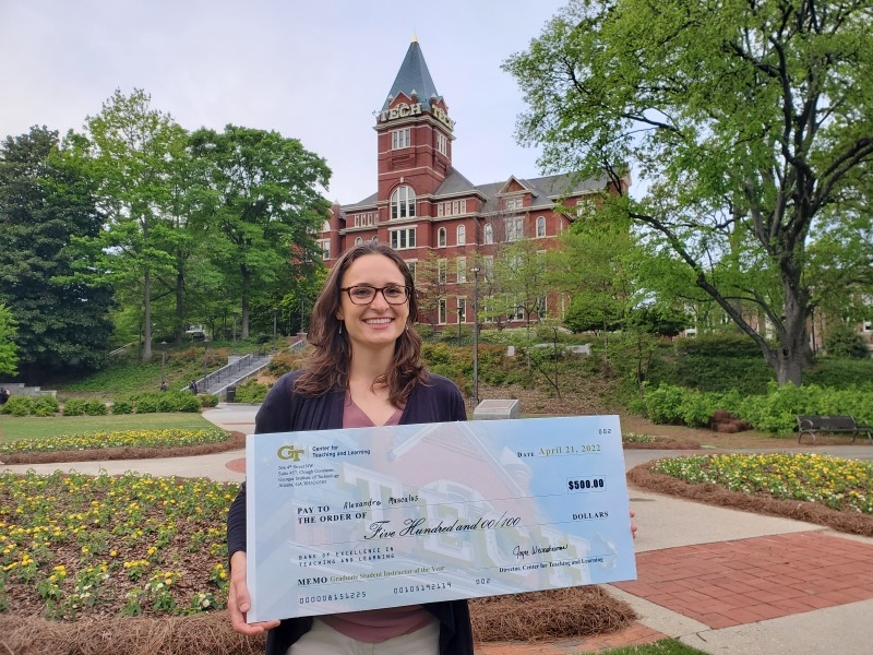 A woman holds a large check in front of a building