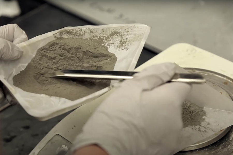 A close up of coal ash and a silver instrument in a dish held by hands in white latex gloves 