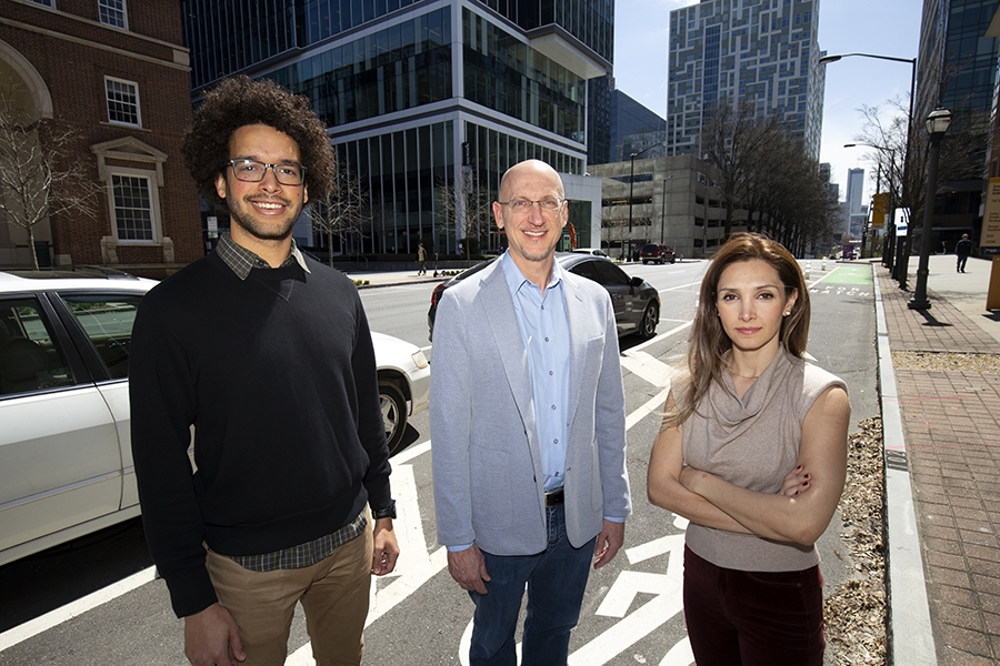 Two men and one woman stand together on the street in front of cars and buildings