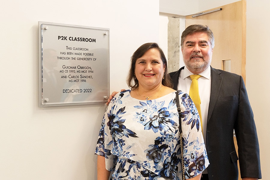 Two people stand next to a silver sign with black text